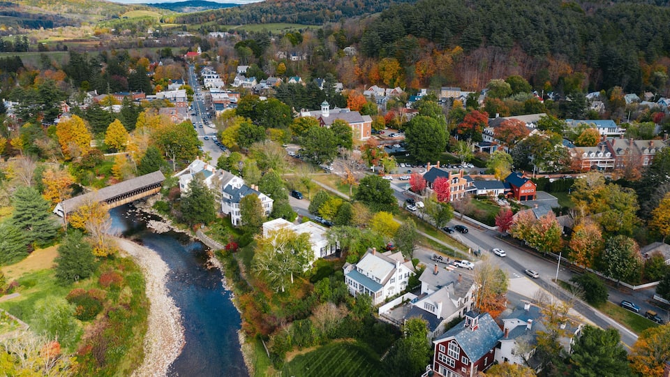 An aerial view of a town with a river, surrounded by autumn-colored trees and a bridge.