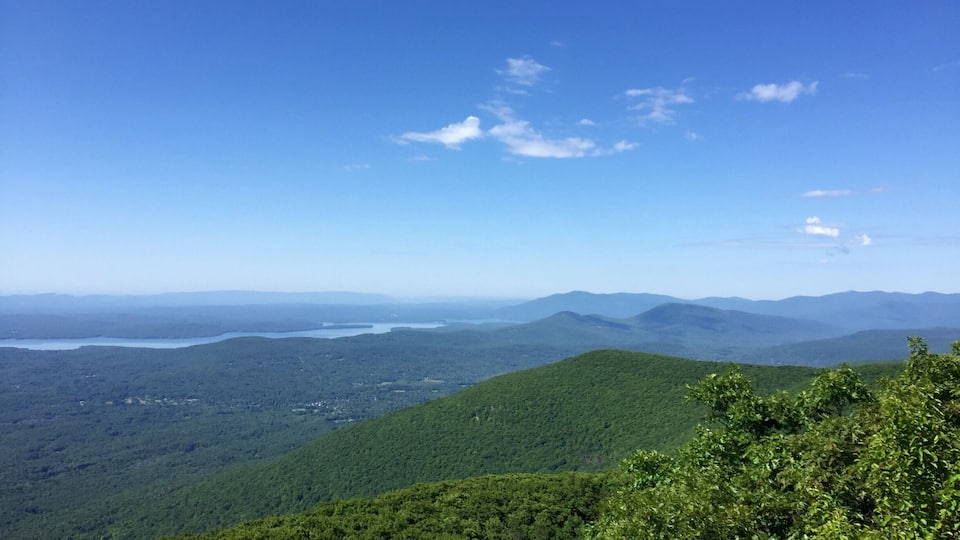A mountainous landscape with dense forest, a lake, and distant mountains under a clear blue sky.