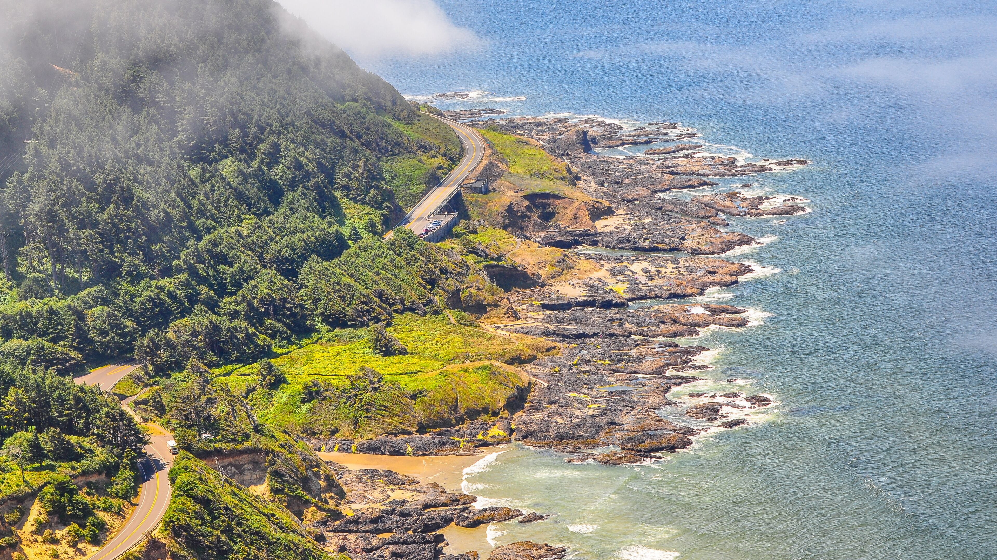 A coastal road winding through a forested area with rocky cliffs and ocean waves.