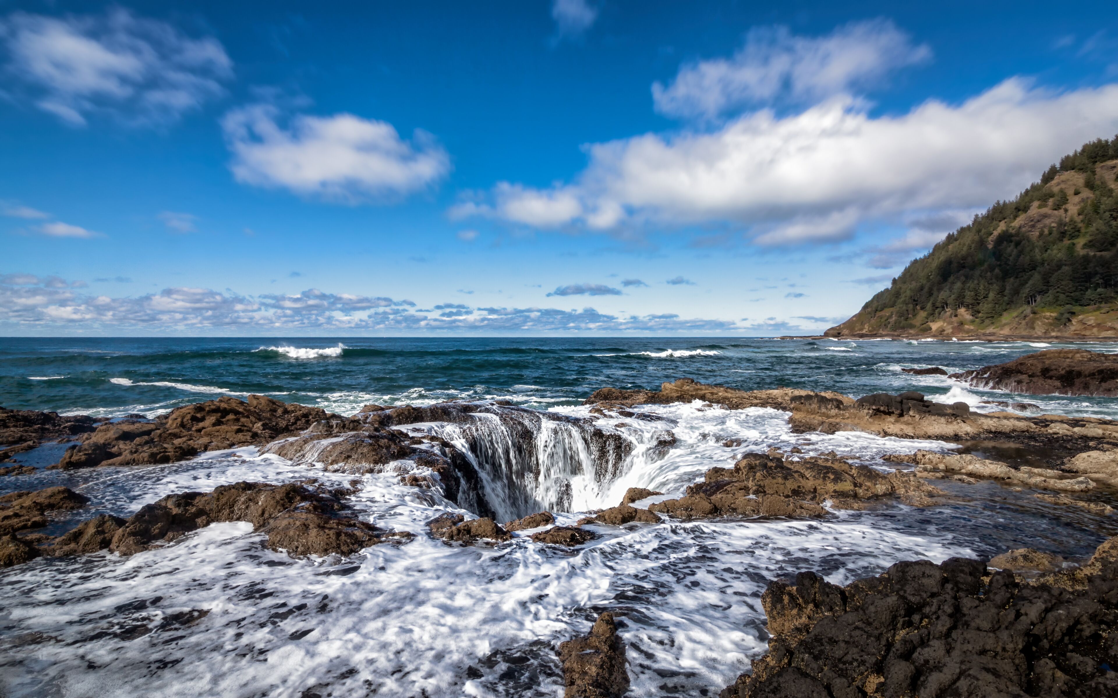 A coastal landscape with a rocky shoreline, waves crashing against the rocks, and a clear blue sky with scattered clouds.