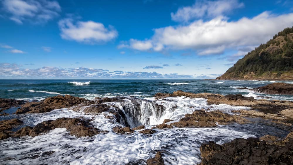 A coastal landscape with a rocky shoreline, waves crashing against the rocks, and a clear blue sky with scattered clouds.