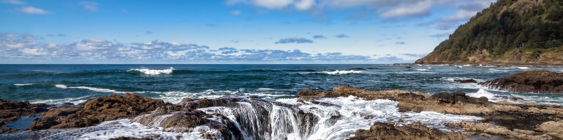 A coastal landscape with a rocky shoreline, waves crashing against the rocks, and a clear blue sky with scattered clouds.