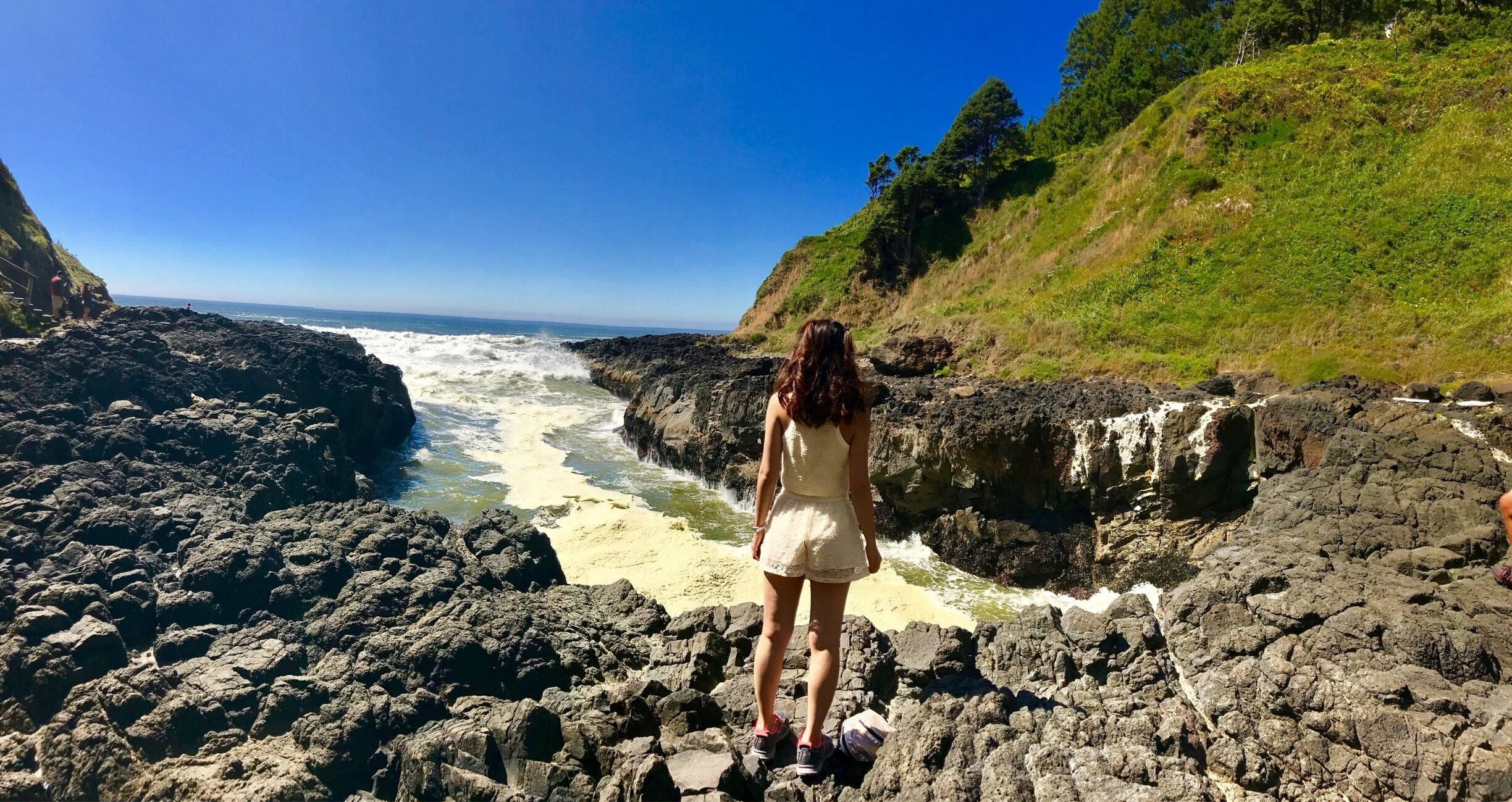 A woman standing on rocky terrain overlooking a coastal area with waves crashing against the rocks.