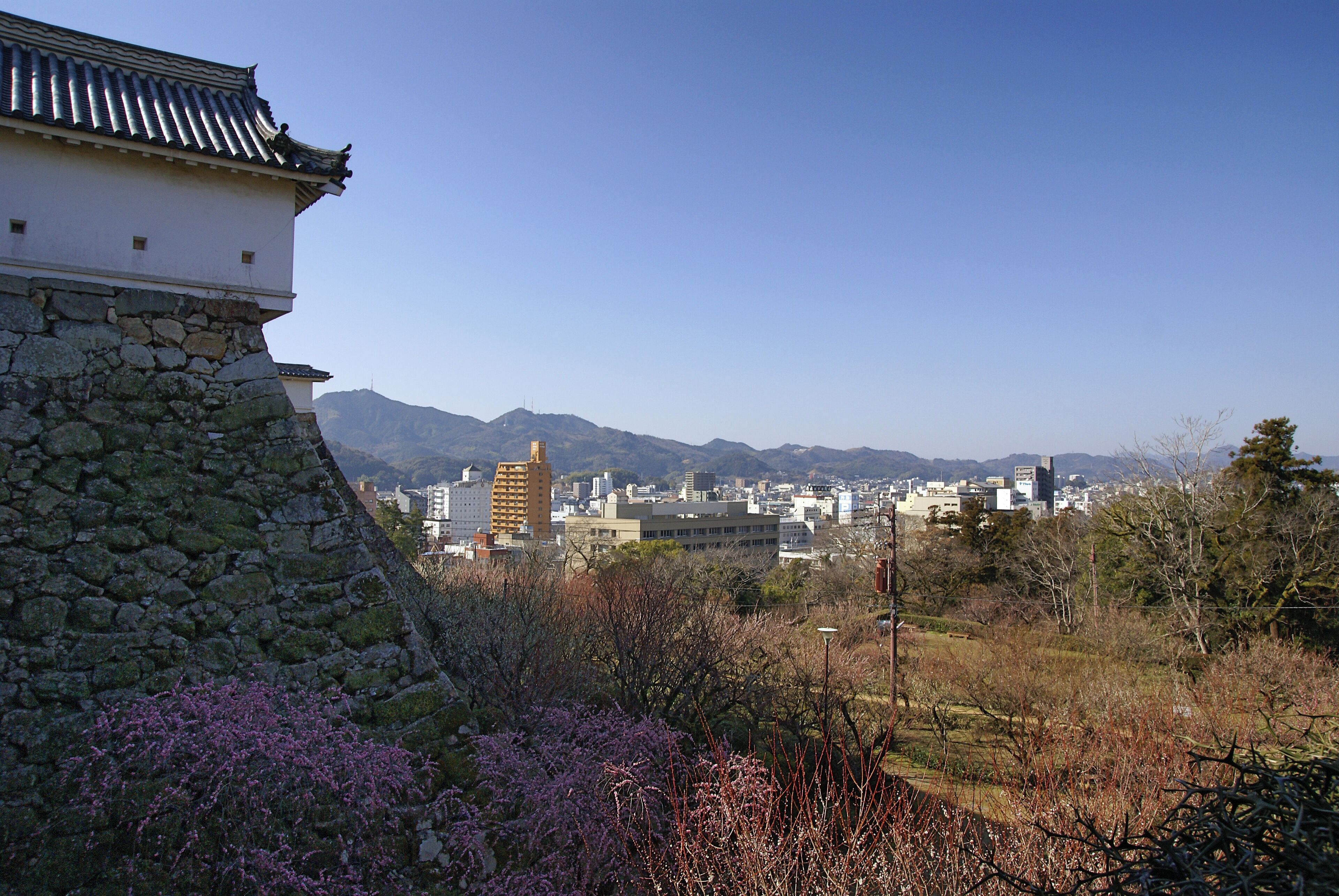 A traditional Japanese building with a stone base and tiled roof, overlooking a cityscape with modern buildings and mountains in the background.