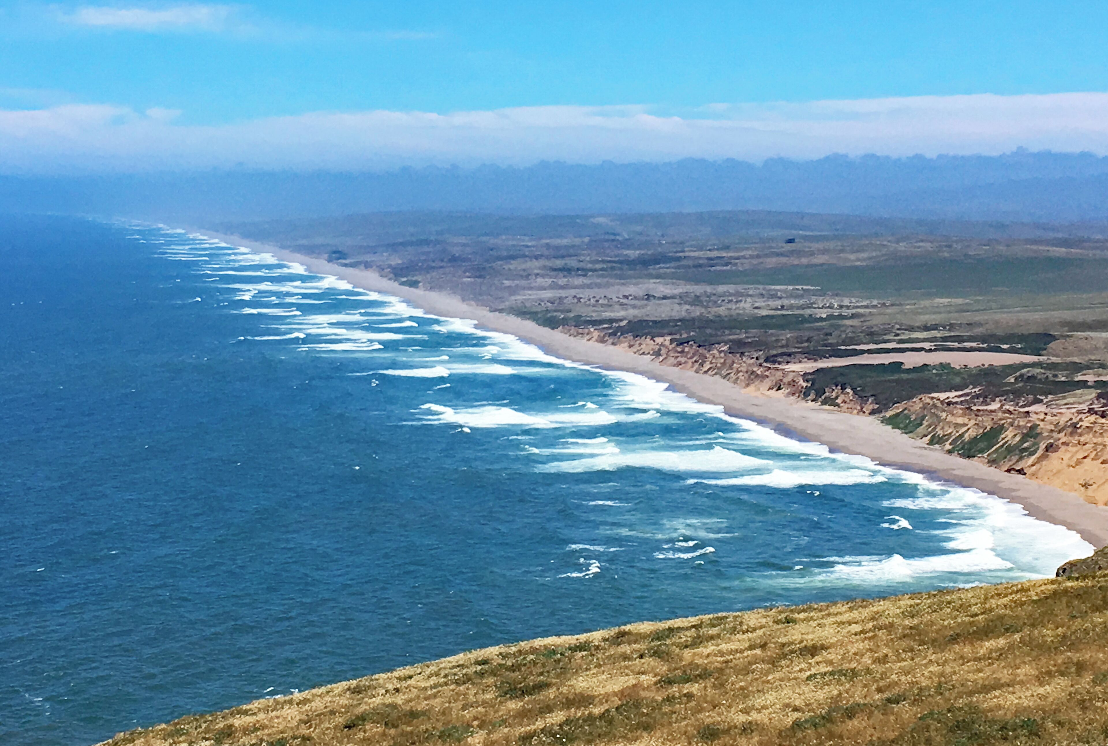 A coastal landscape with a long stretch of beach, ocean waves, and distant mountains.