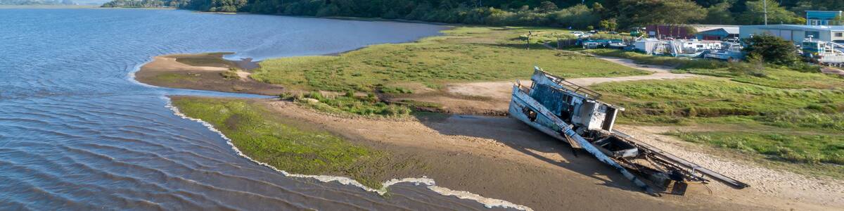 Aerial view of a shipwrecked boat on a sandy beach in Inverness, California, USA. Two people walk nearby, observing the wreckage. The boat is old and decaying.
