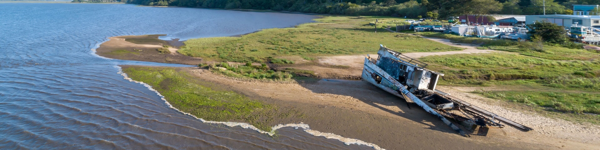 Aerial view of a shipwrecked boat on a sandy beach in Inverness, California, USA. Two people walk nearby, observing the wreckage. The boat is old and decaying.