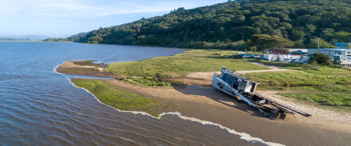 Aerial view of a shipwrecked boat on a sandy beach in Inverness, California, USA. Two people walk nearby, observing the wreckage. The boat is old and decaying.
