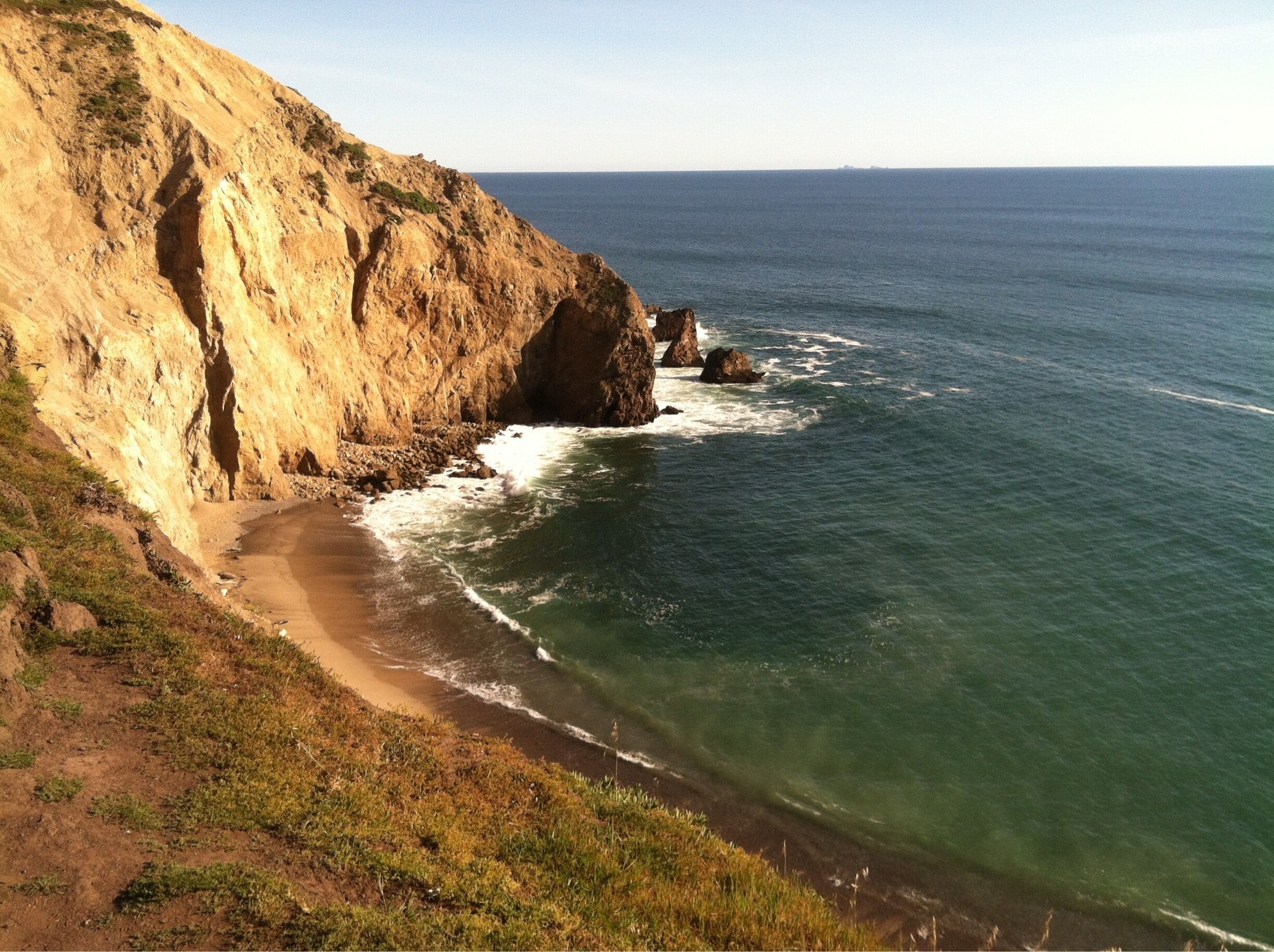 A coastal cliff with a sandy beach and clear blue sea.