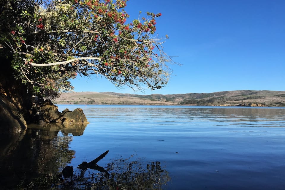 A calm lake with clear reflections, a rocky shoreline, and distant hills.