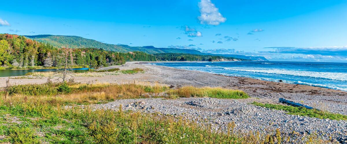 A coastal landscape with a rocky beach, a calm lake, and a forested hillside.