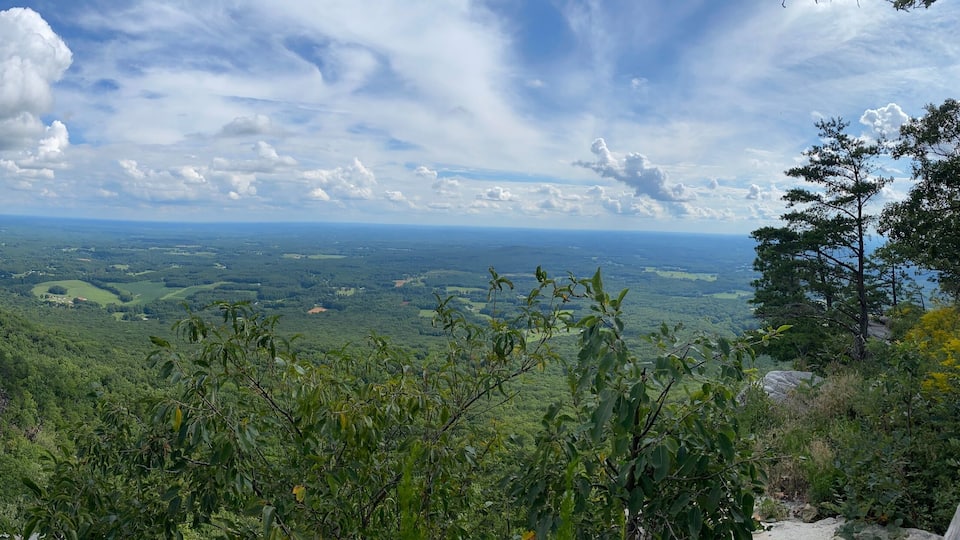 A mountainous landscape with dense forests, rocky outcrops, and a distant view of a valley under a partly cloudy sky.