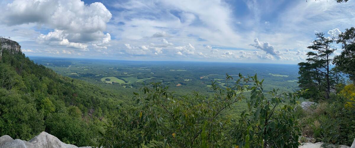 A mountainous landscape with dense forests, rocky outcrops, and a distant view of a valley under a partly cloudy sky.