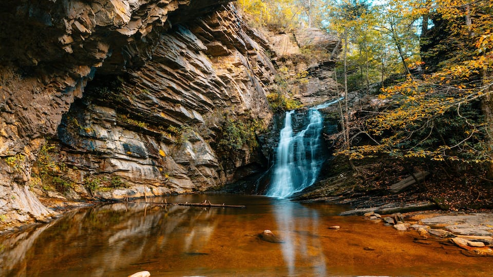 A waterfall cascading into a clear pool surrounded by rocky cliffs and autumn foliage.