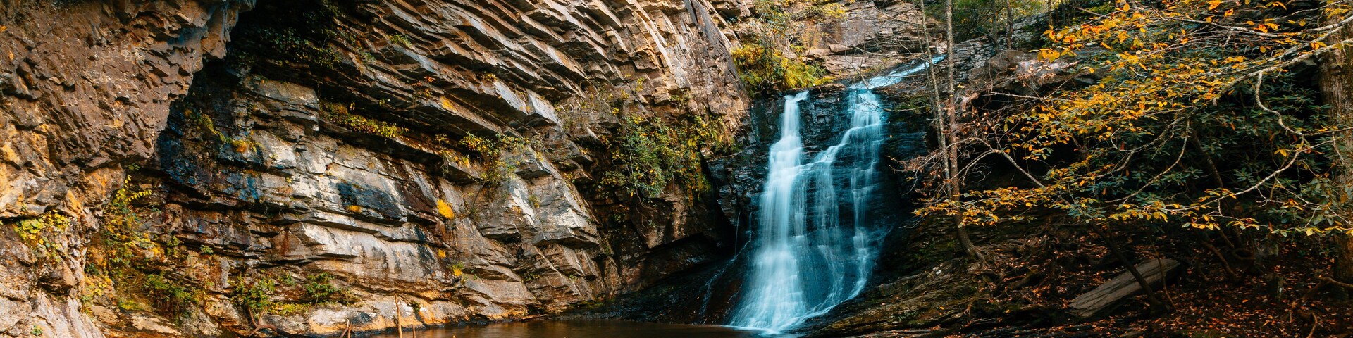 Una cascada se precipita en una piscina cristalina rodeada de riscos y follaje otoñal.