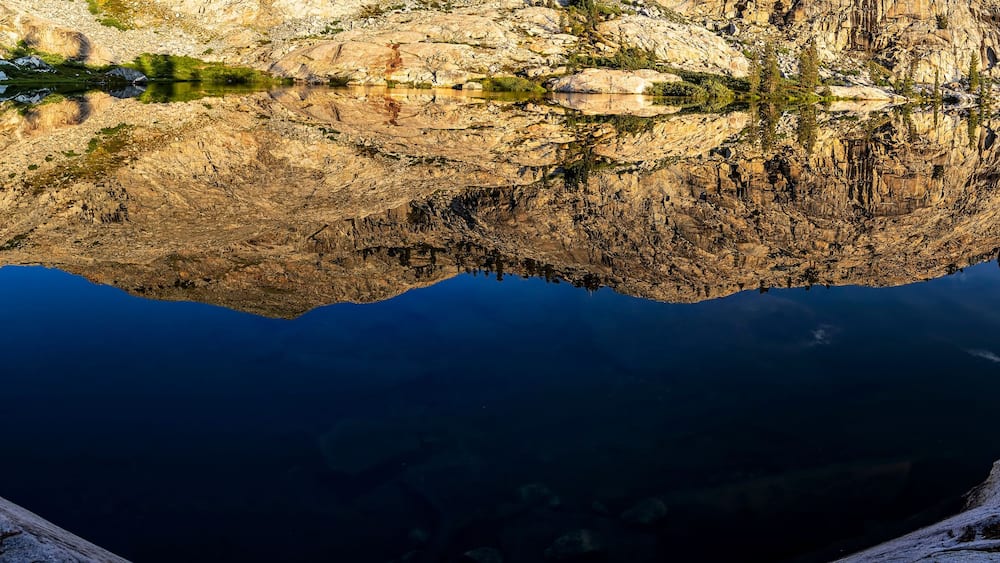 A mountain lake with clear reflections of the surrounding rocky terrain and sparse vegetation.