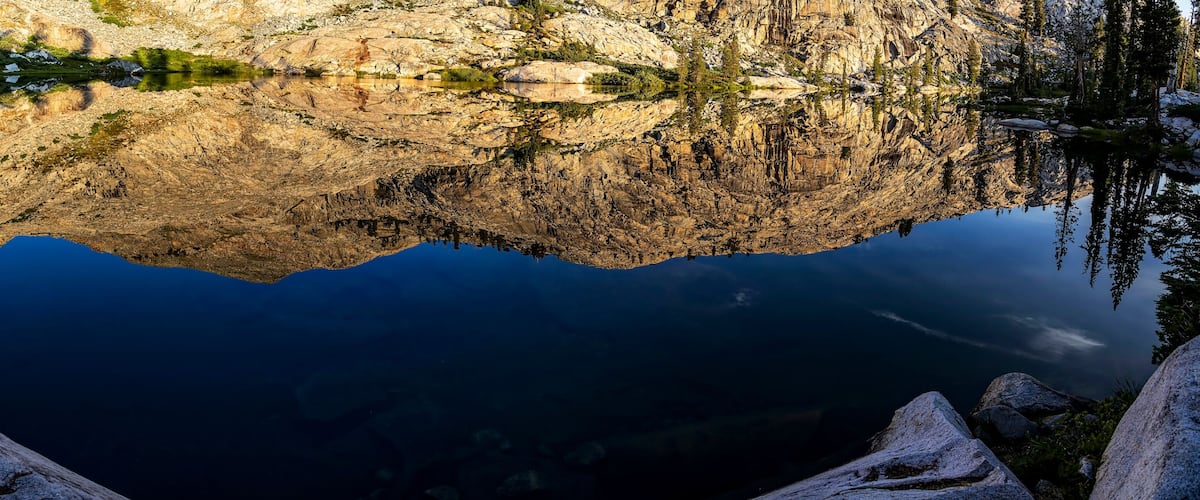 A mountain lake with clear reflections of the surrounding rocky terrain and sparse vegetation.