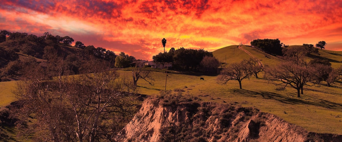 A sunset over a hilly landscape with sparse trees and a clear sky.