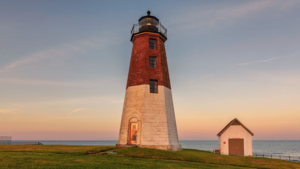 A lighthouse with a red and white tower, a small adjacent building, and a grassy area with a view of the sea.
