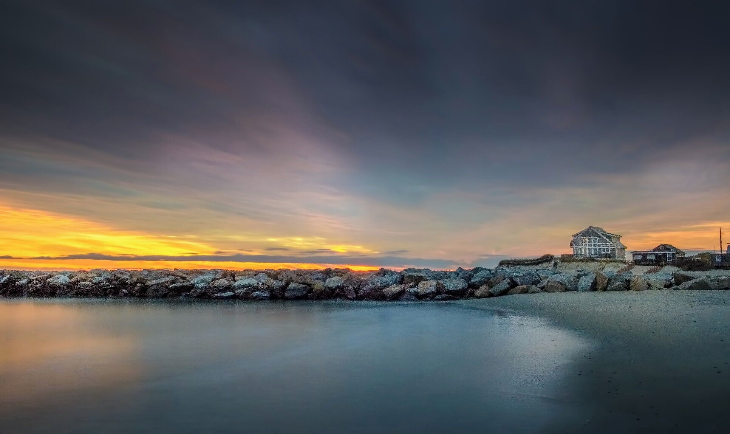 Paisaje costero con un rompeolas rocoso, una casa blanca y un atardecer.