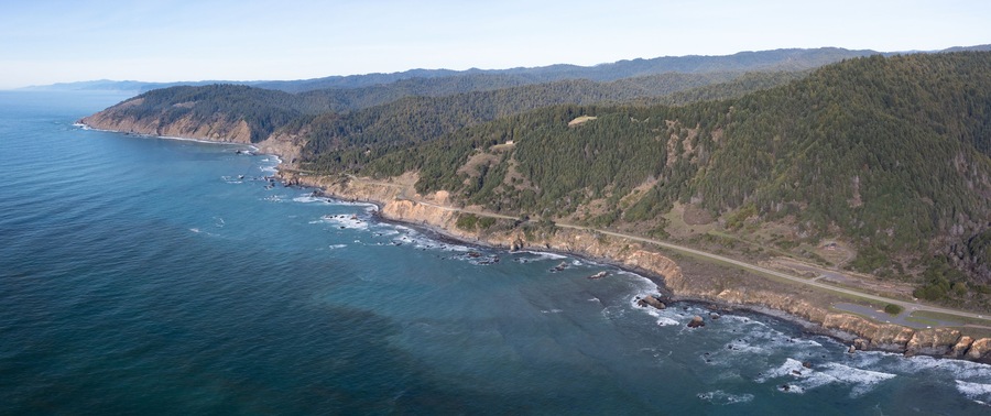 A coastal landscape with a road along the cliffside, rocky shoreline, and dense forested hills.