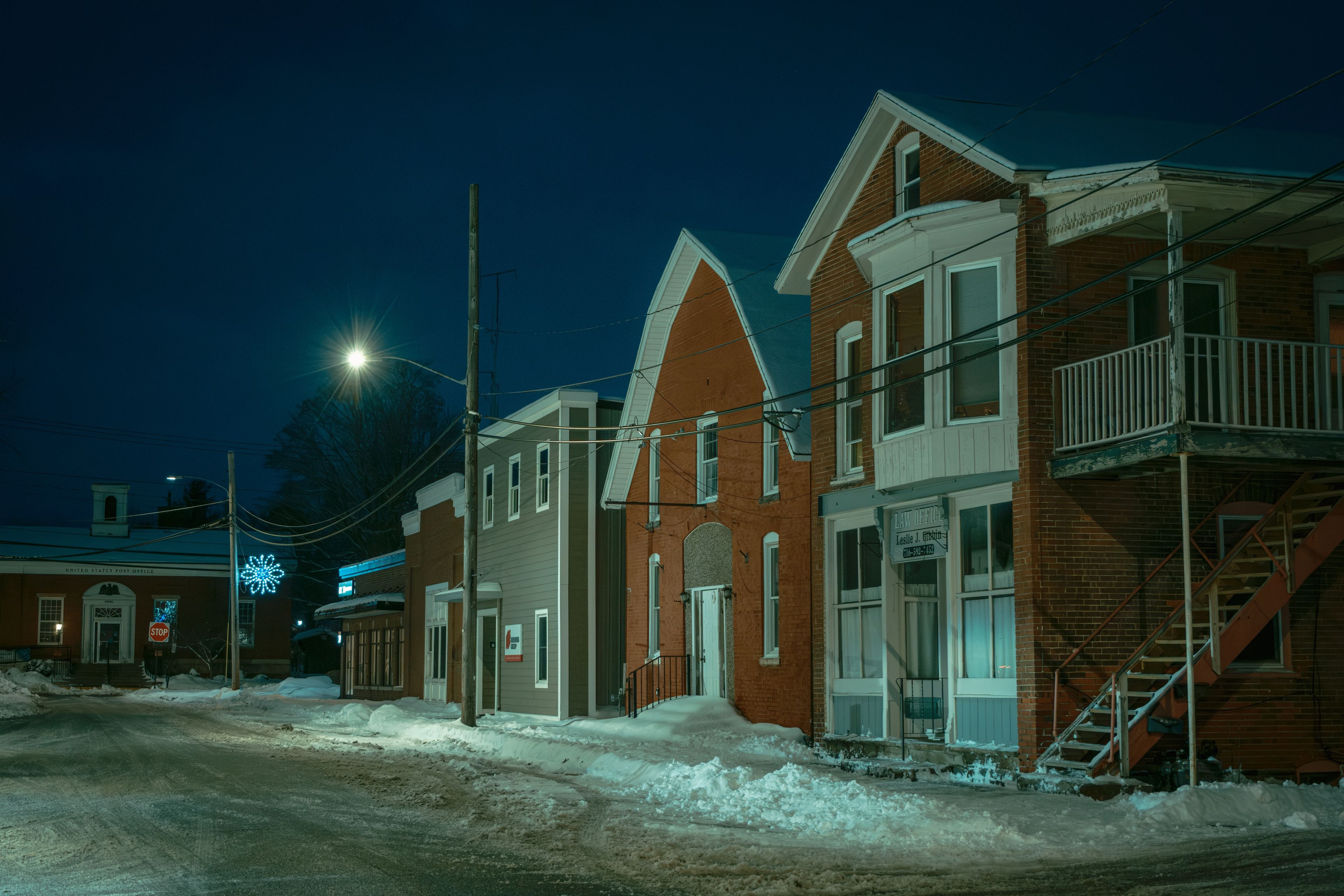 Eine verschneite Straße bei Nacht mit Ziegelgebäuden, einem Stoppschild und einem United States Post Office.