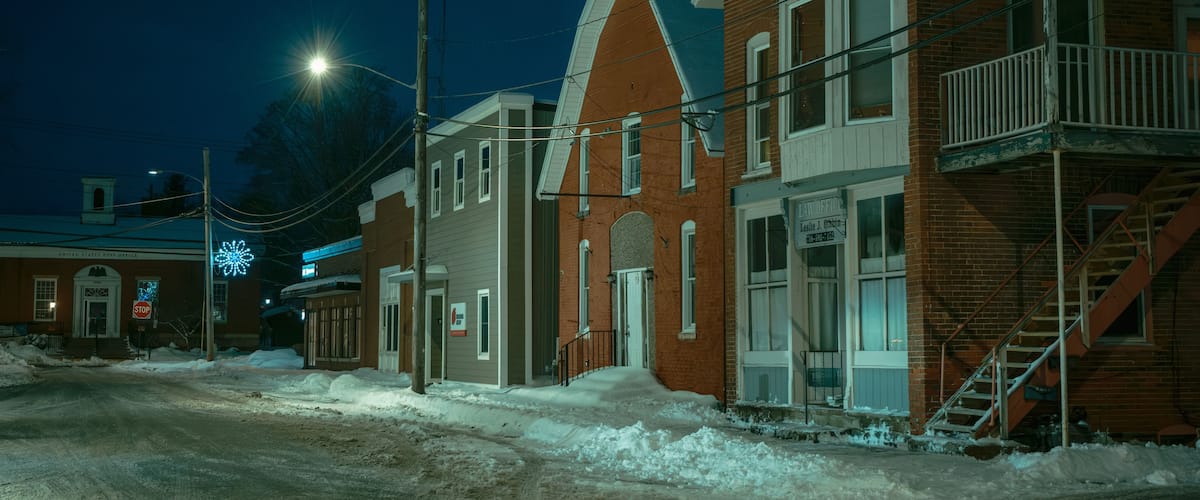 A snow-covered street at night with brick buildings, a stop sign, and a United States Post Office.