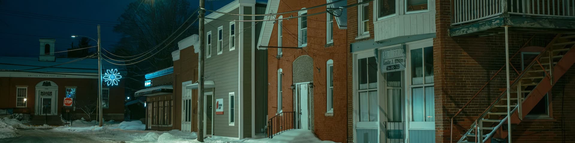 A snow-covered street at night with brick buildings, a stop sign, and a United States Post Office.