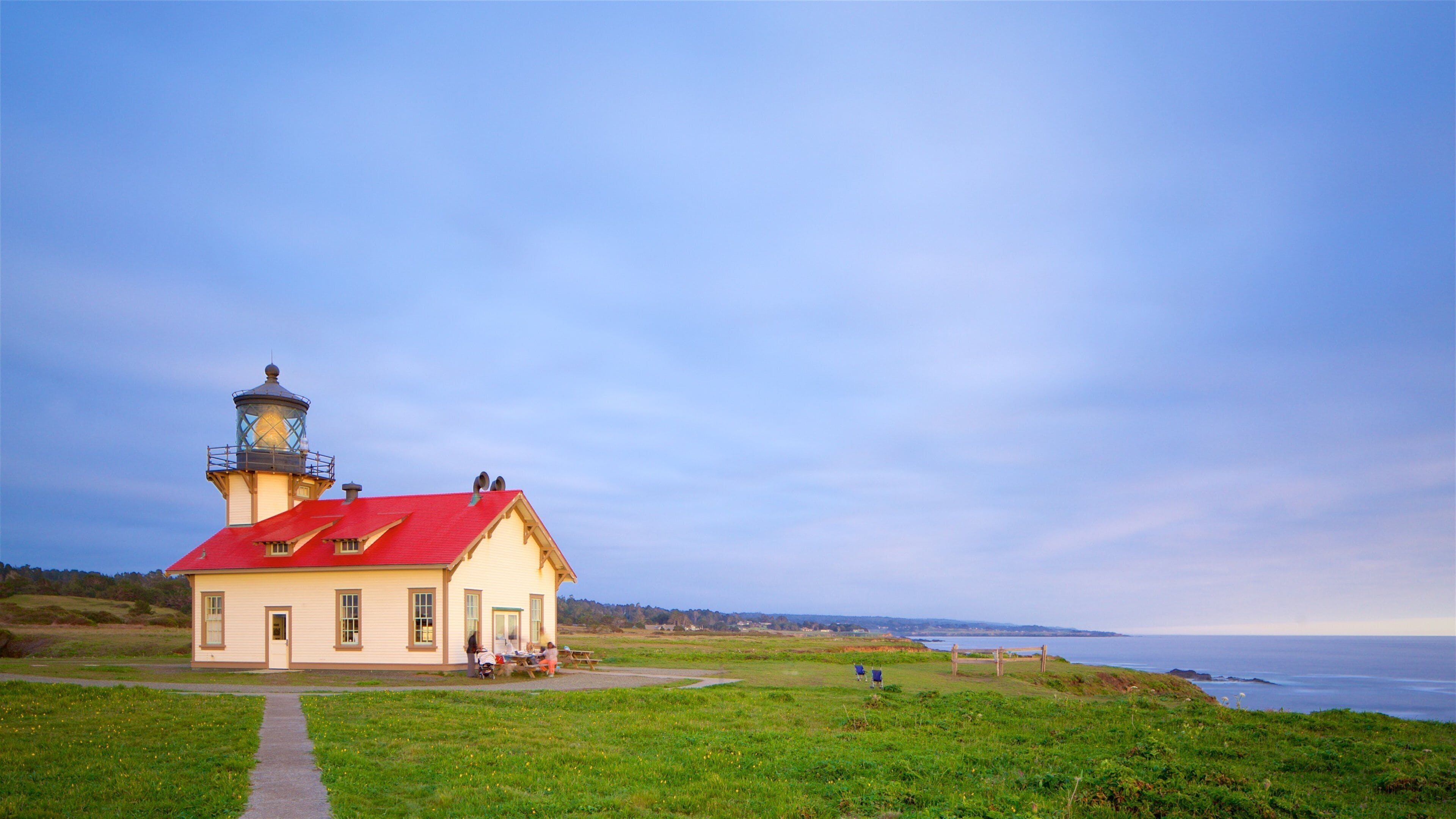A lighthouse with a red roof and a white building, situated on a grassy hill overlooking the ocean.