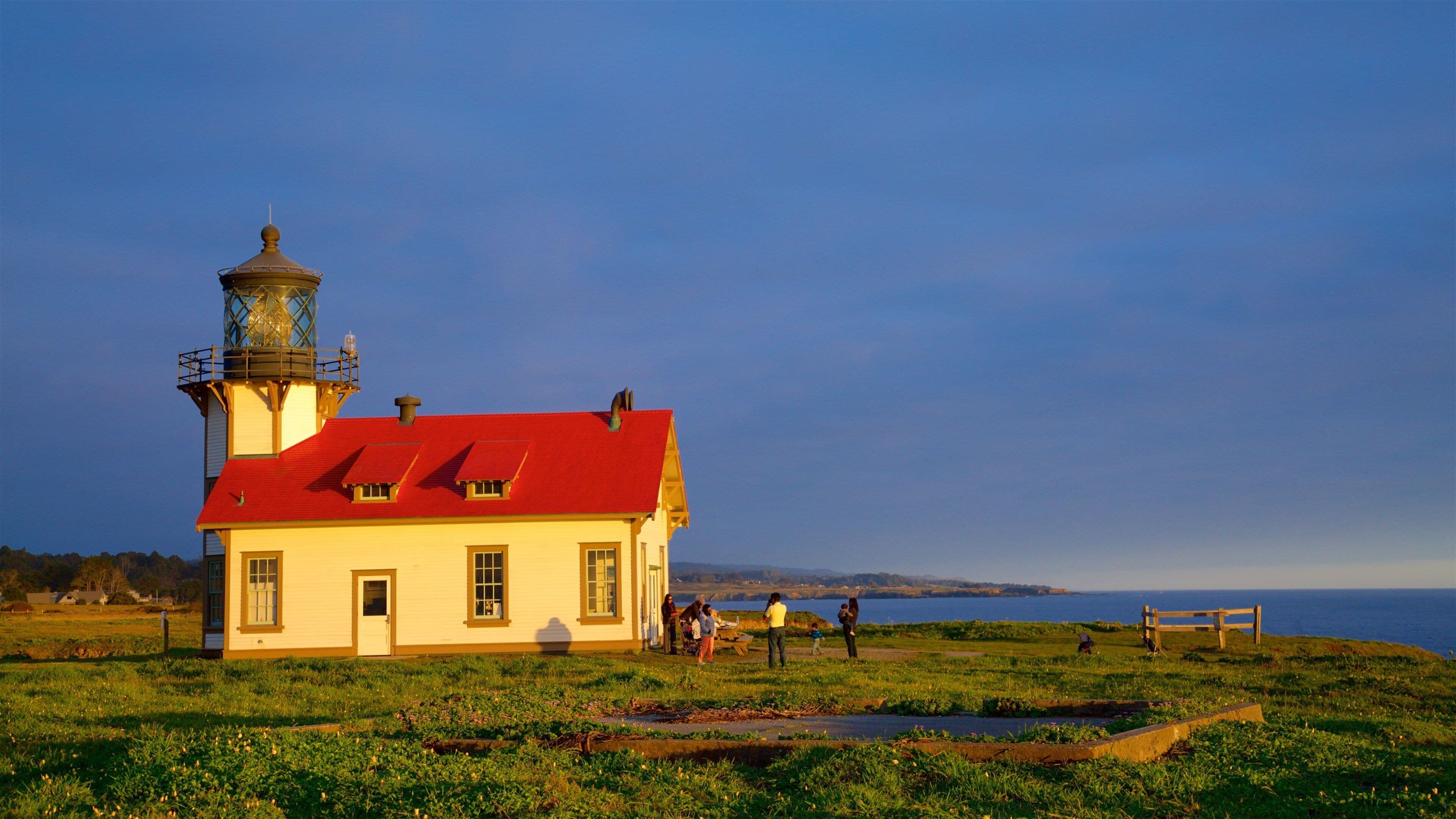 A lighthouse with a red roof and a tower, surrounded by grassy fields and a body of water.