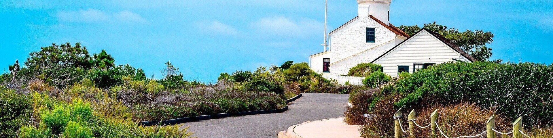 Um farol branco com topo verde, cercado por vegetação e uma estrada sinuosa.