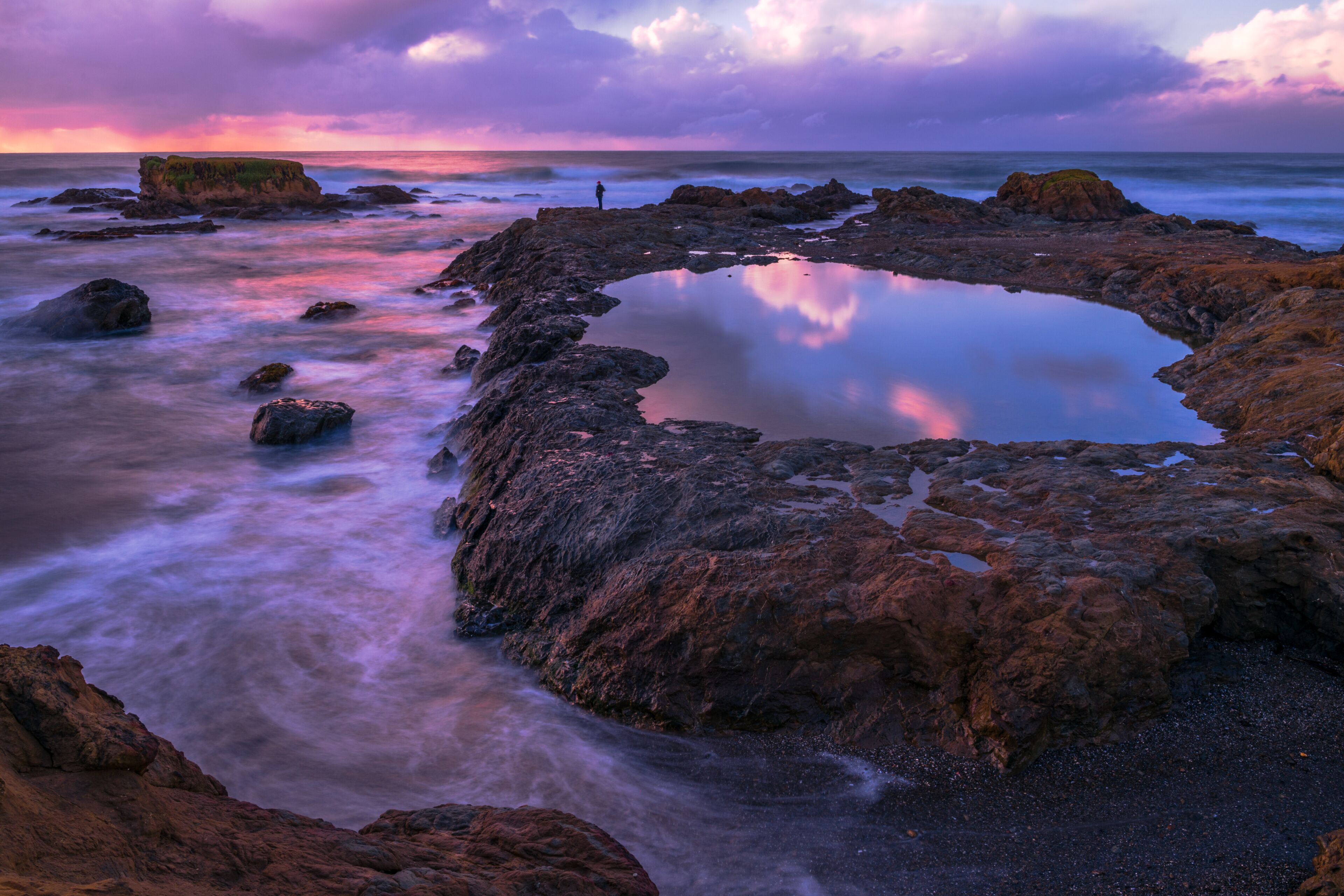 A rocky shoreline with a reflective pool and a person standing on the rocks.