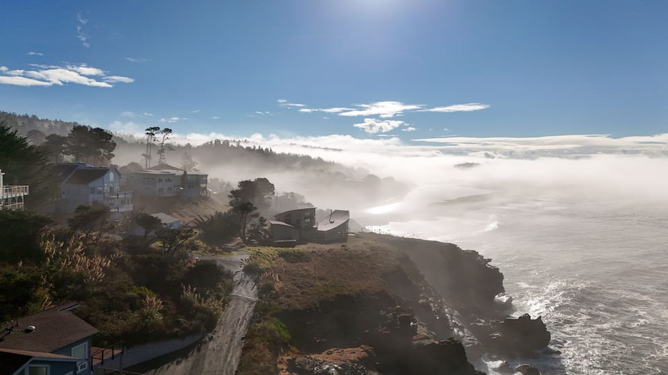 A coastal landscape with houses on a cliff overlooking the ocean, surrounded by fog.