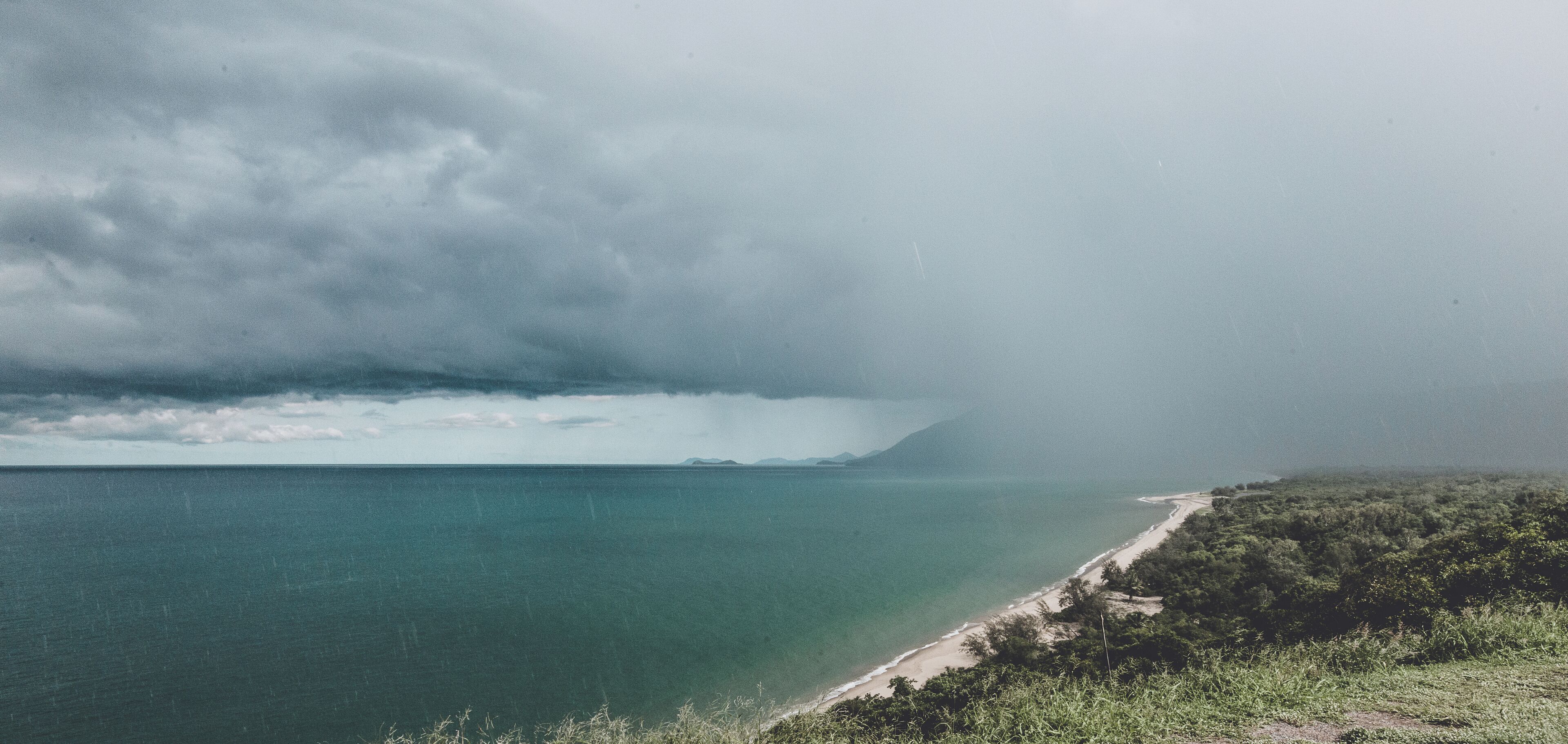 A coastal landscape with a beach, dense vegetation, and a stormy sky.