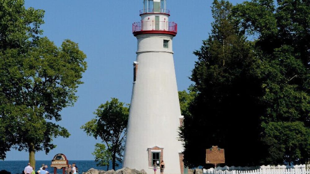 A white lighthouse with a red top, surrounded by trees and a white picket fence.