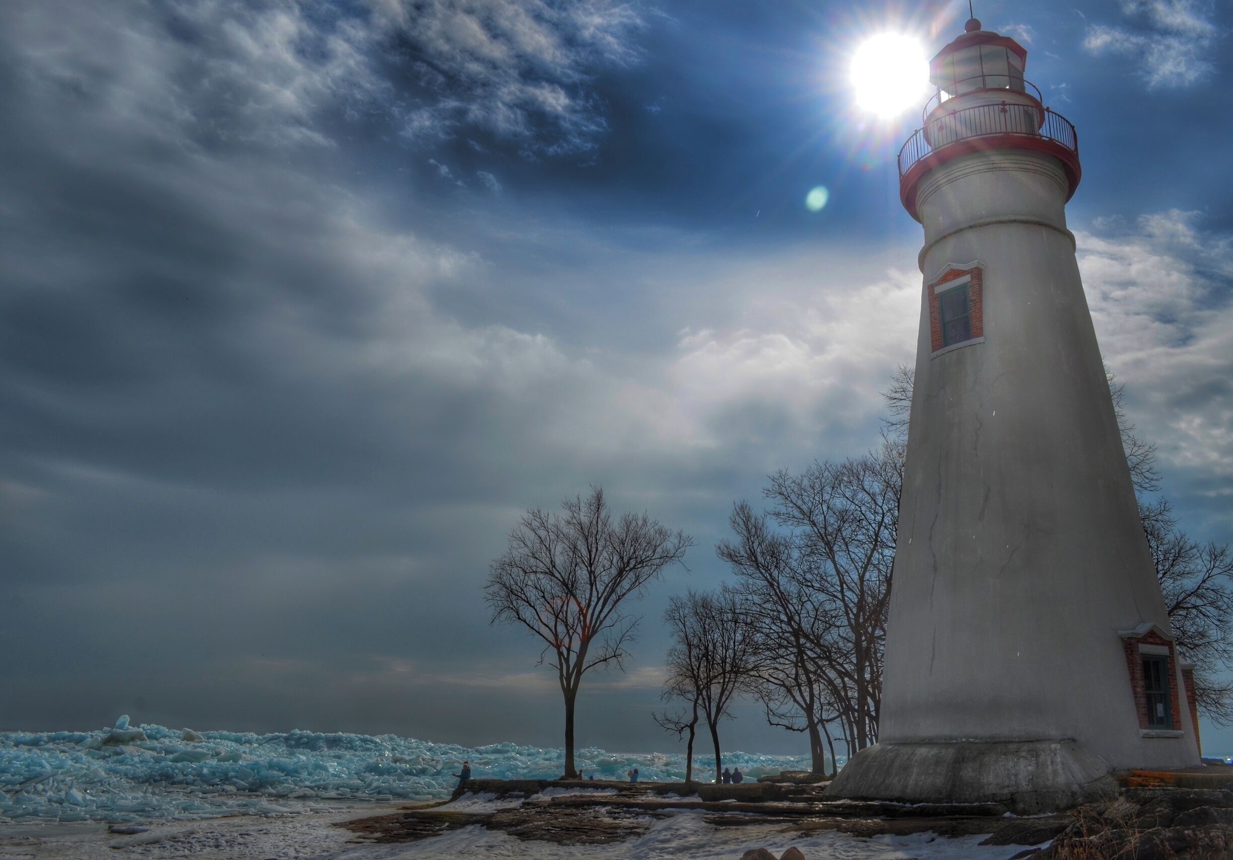 A lighthouse with a red top, surrounded by bare trees and a snowy landscape under a cloudy sky.