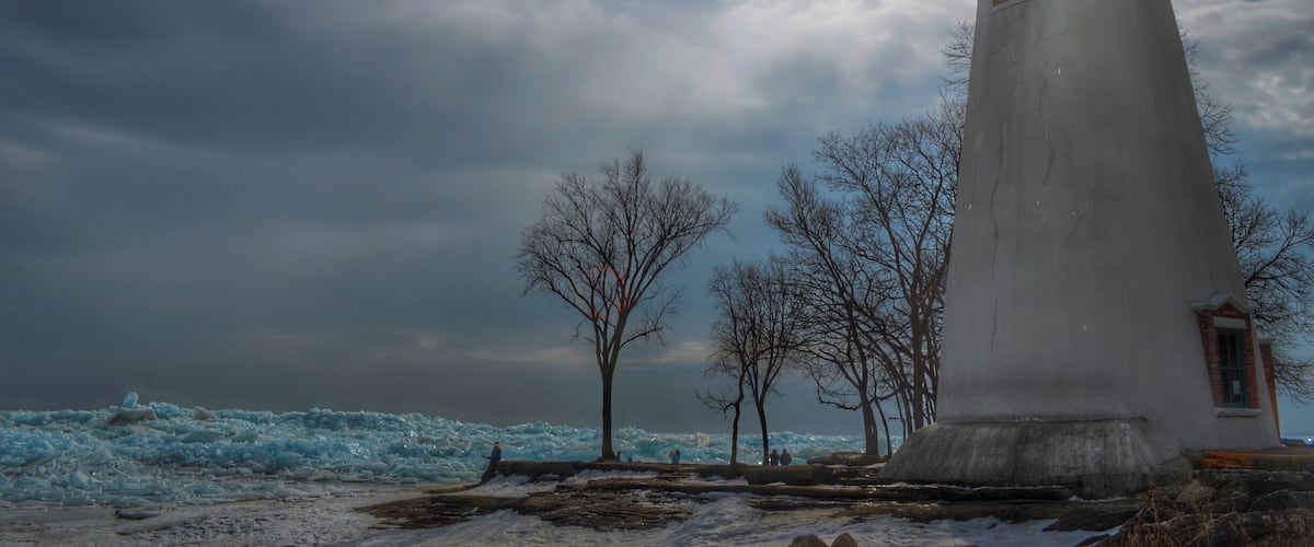 A lighthouse with a red top, surrounded by bare trees and a snowy landscape under a cloudy sky.
