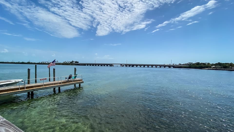 A clear waterfront with a wooden dock, an American flag, and a bridge in the background.