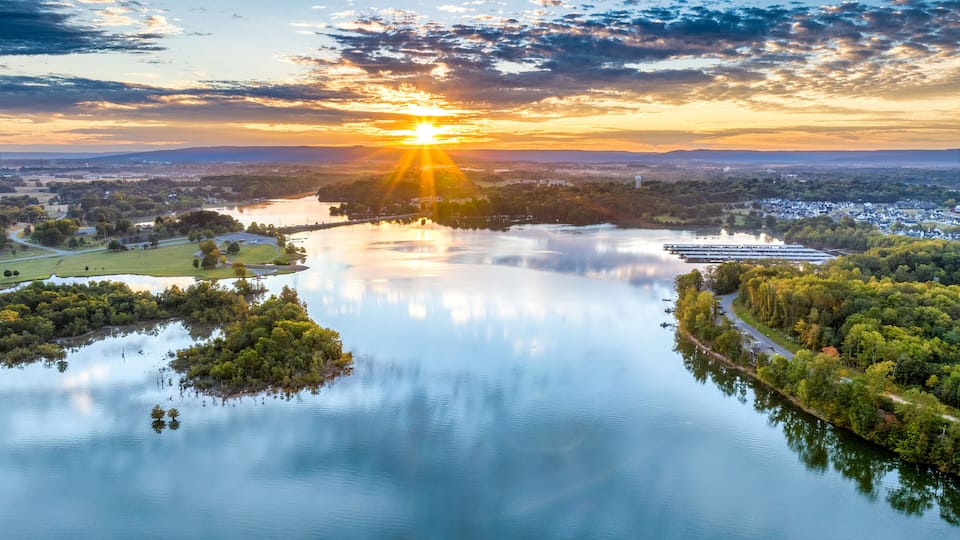 A river reflecting the sunset, surrounded by greenery and a distant cityscape.