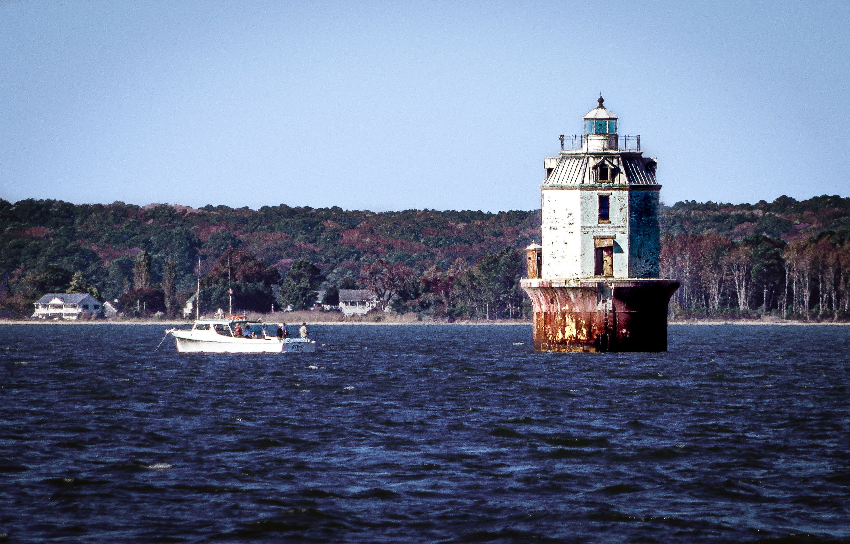 A lighthouse on a rocky outcrop surrounded by water, with a boat in the foreground and a forested shoreline in the background.