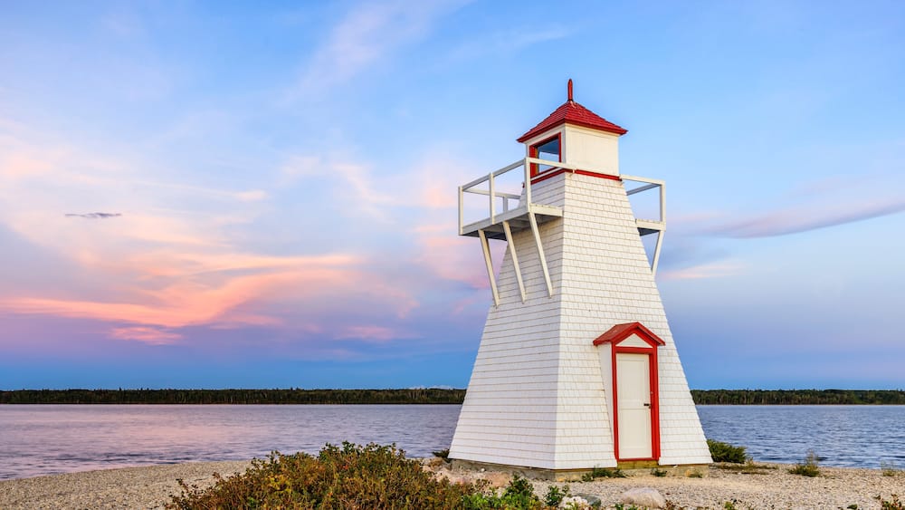 A white lighthouse with a red roof and door, situated on a rocky shoreline with a calm lake and a forested background.