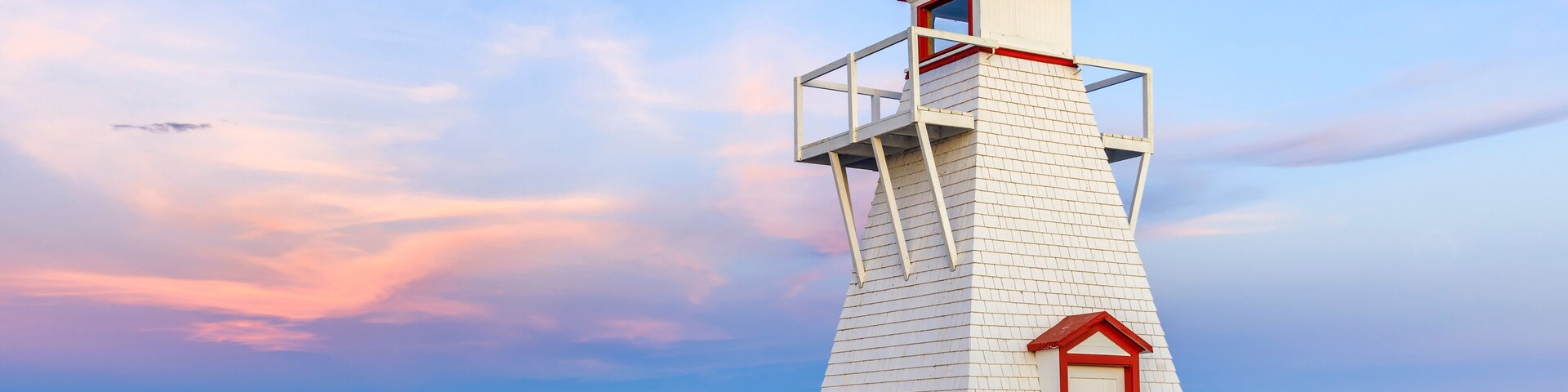 Um farol branco com teto e porta vermelhos, localizado numa costa rochosa com um lago calmo e uma área verde ao fundo.