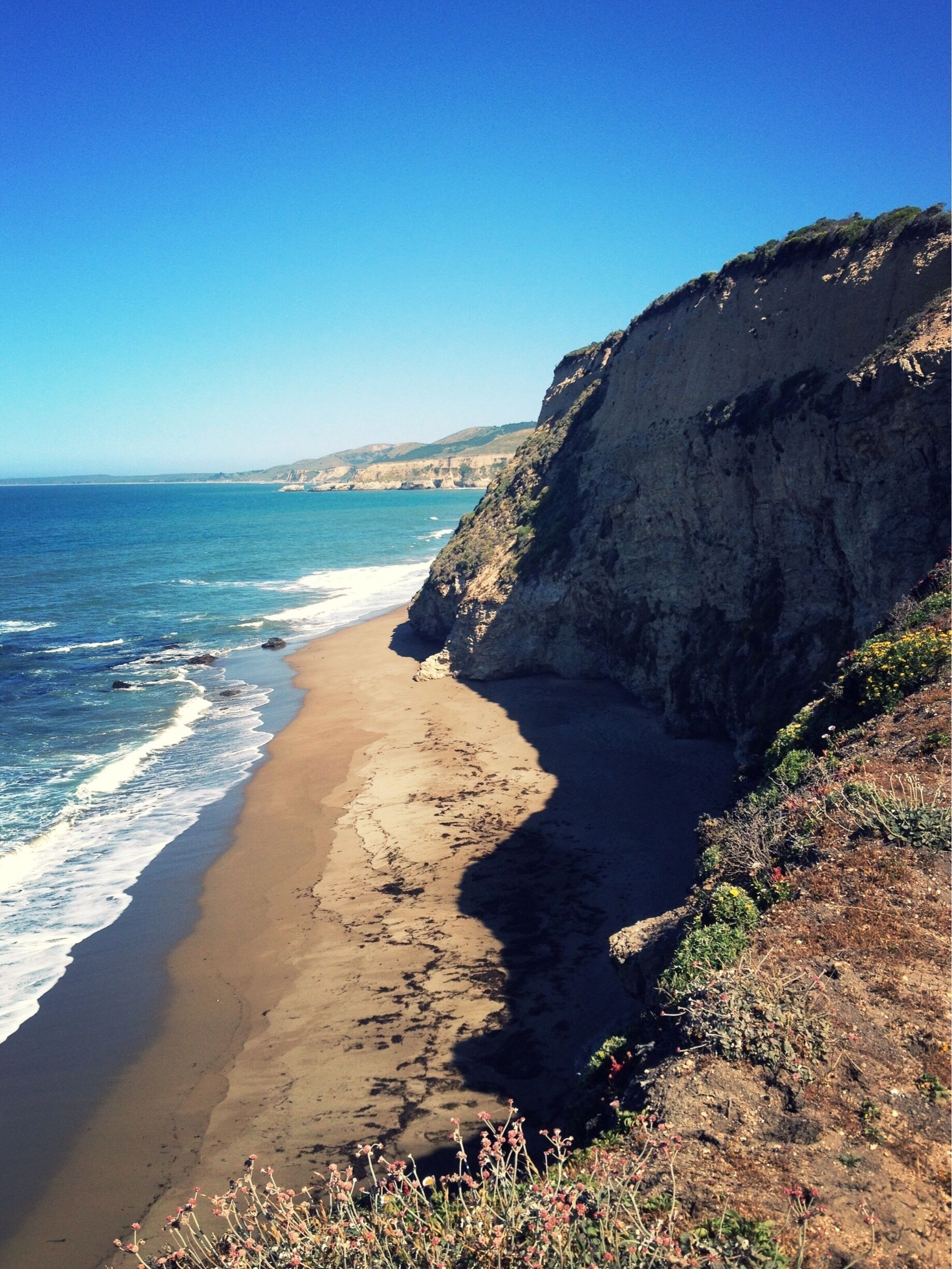 A coastal landscape with a sandy beach, ocean waves, and a steep cliff.