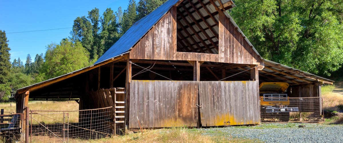 A rustic wooden barn with a slanted roof, surrounded by a wire fence and a gravel path.