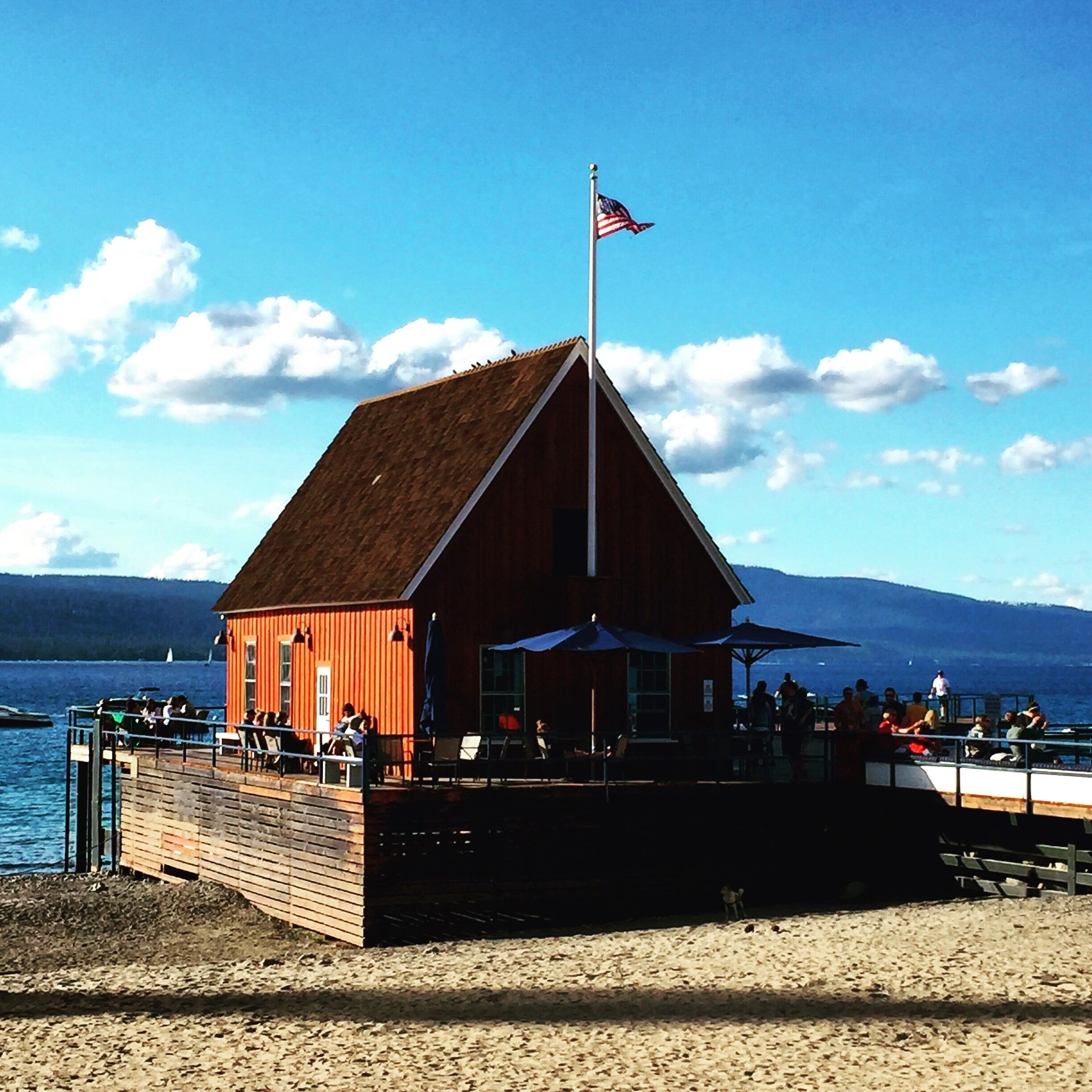 A wooden building with a red exterior and a flagpole, located on a beach with people sitting outside.