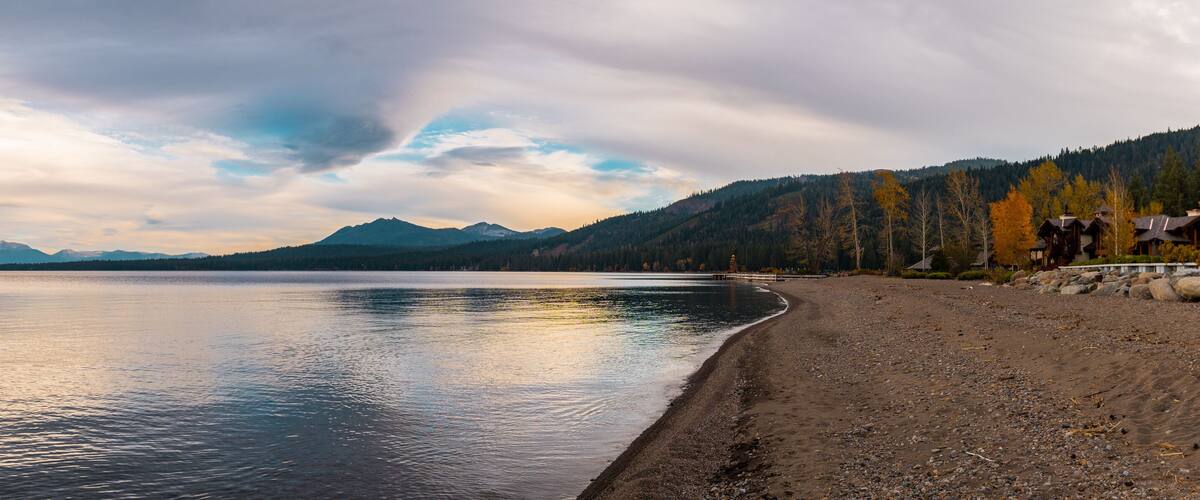 A calm lake with a rocky shoreline, surrounded by mountains and autumn-colored trees.