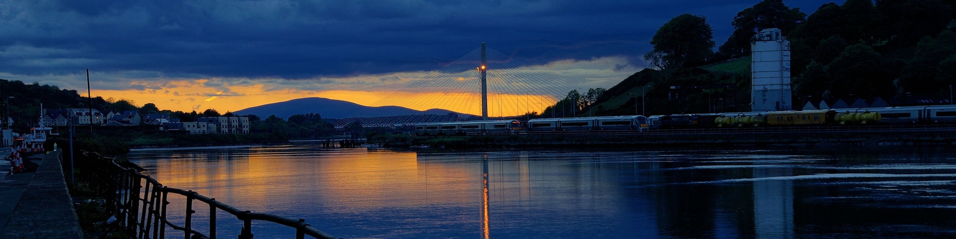 Un río al atardecer con una cerca de madera, un puente y una cadena montañosa distante.