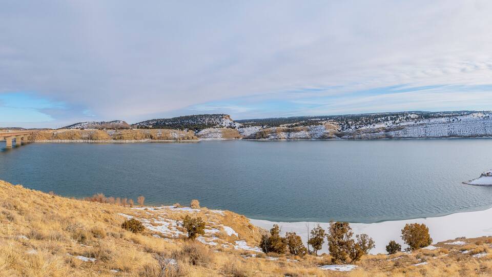 A snow-covered landscape with a bridge over a calm lake, surrounded by hills and sparse vegetation.
