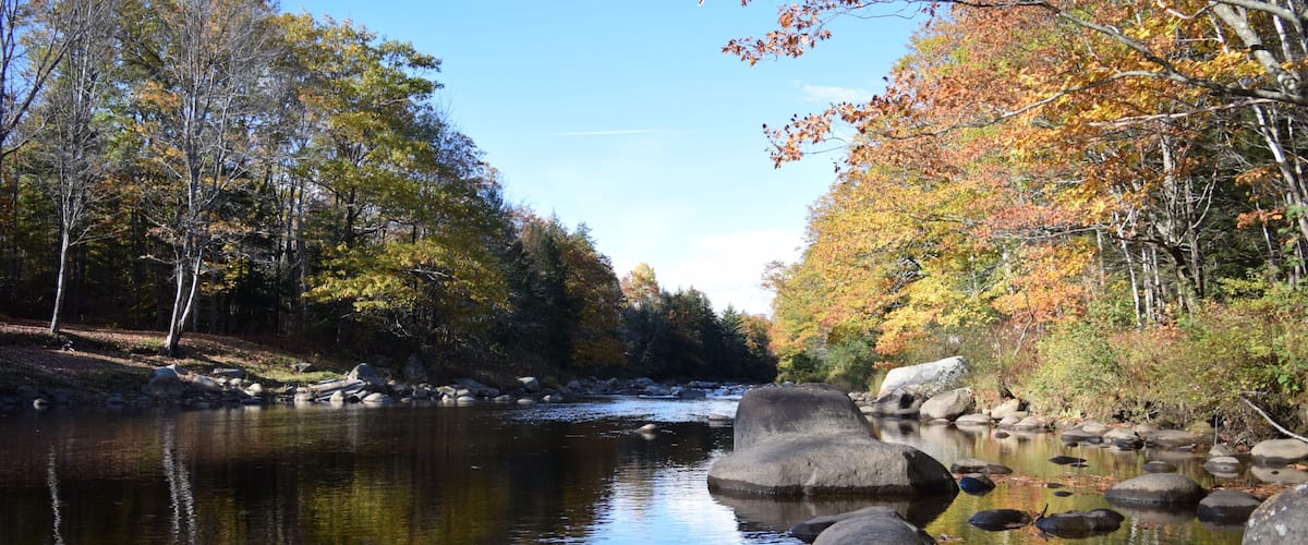 A calm river with large rocks, surrounded by trees with autumn foliage.