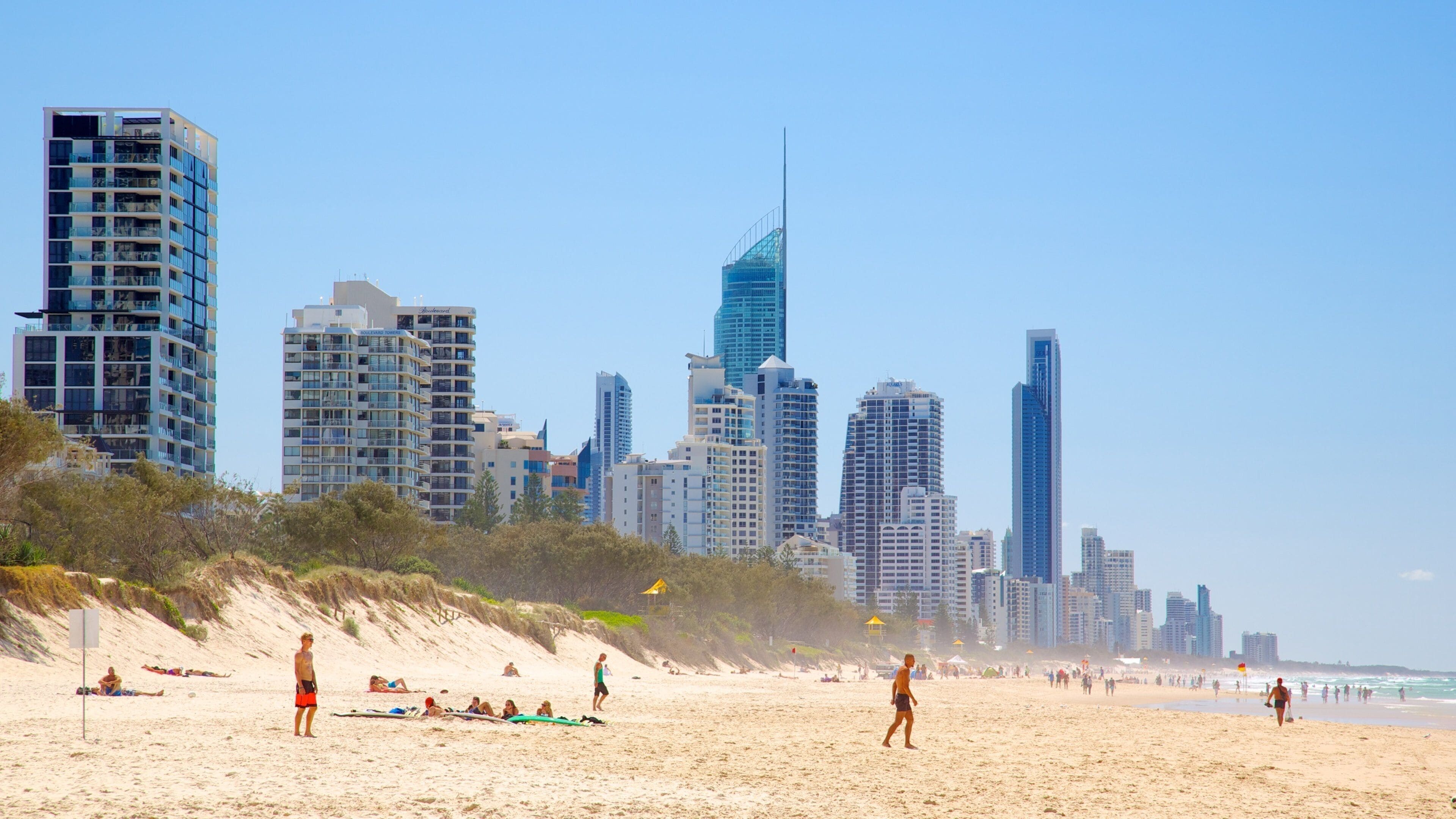 Una playa con gente tomando el sol y nadando, y un horizonte urbano con edificios altos al fondo.